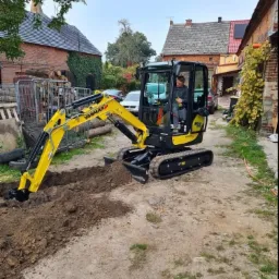 Żółta mini koparka Yanmar kopie wąski rów w ziemi na tle zabudowań z czerwonej cegły i zaparkowanych samochodów.