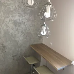 Ściana wykończona dekoracyjnym tynkiem w odcieniach szarości, oświetlona trzema wiszącymi lampami w metalowych kloszach, z drewnianym blatem i dwoma hokerami.