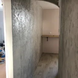 Korytarz z dekoracyjną, szarą ścianą o nieregularnej fakturze i podłogą imitującą drewno, widok na wnękę z półką i gniazdkiem elektrycznym.