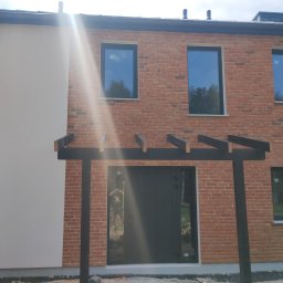 Usługi Remontowo-Budowlane - Dwukondygnacyjny dom z elewacją z czerwonej cegły i fragmentem ściany otynkowanej na biało, z czarnymi oknami i pergolą nad wejściem, w słoneczny dzień.