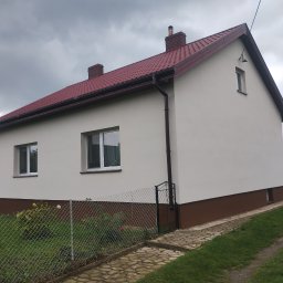 Usługi Remontowo-Budowlane - Dom jednorodzinny po remoncie elewacji, z nową, jasną elewacją i ciemnobrązowym cokołem. Czerwony dach, widoczne okna i fragment ogrodzenia.