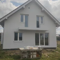 Dwukondygnacyjny dom jednorodzinny z szarą elewacją, brązowym dachem i białymi oknami, na pierwszym planie drewniany stół z puszkami farby.