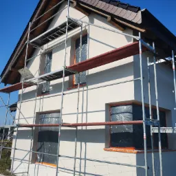 Dom jednorodzinny w trakcie ocieplania elewacji, widoczne rusztowania, okna zabezpieczone folią, świeżo nałożony tynk, brązowa dachówka.