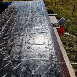 Czarna folia izolacyjna z nadrukiem IZO-HAN Nexler, rozłożona na drewnianej konstrukcji dachu w trakcie budowy, widok z góry, w tle zielona trawa i drzewa.