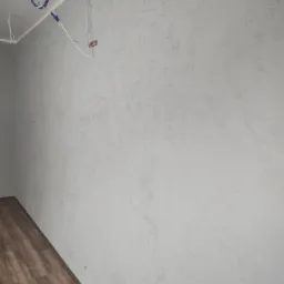 Ściana wewnętrzna pokryta dekoracyjną, jasnoszarą fakturą, widoczne wystające kable elektryczne i fragment drewnianej podłogi.