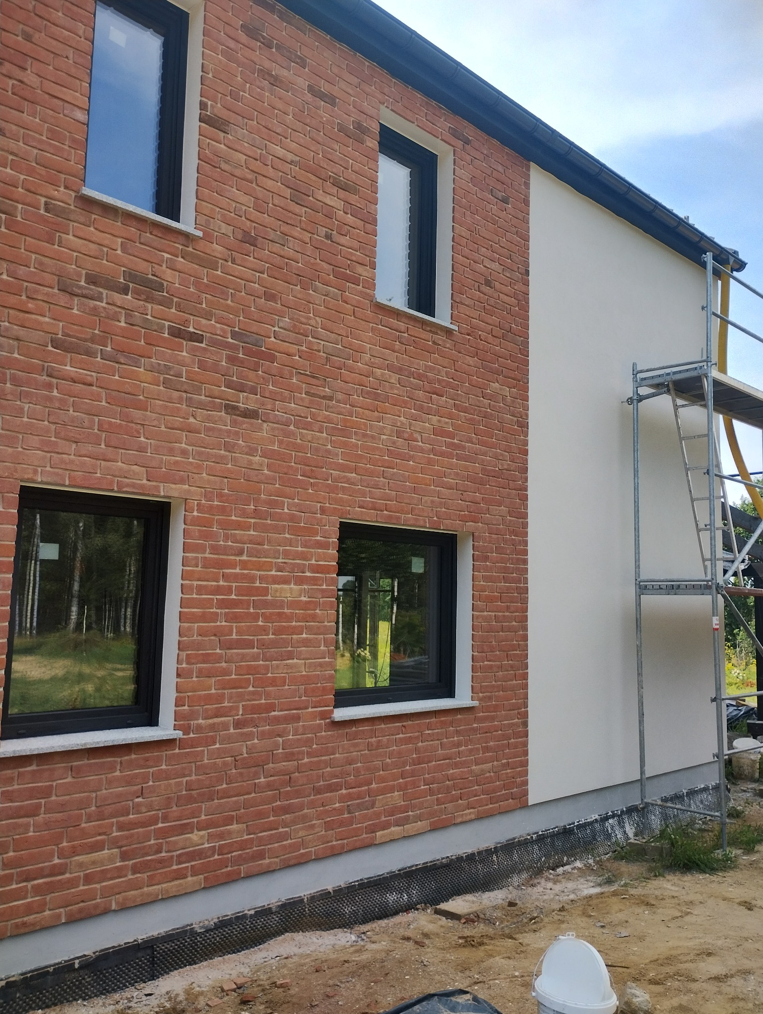 Fragment elewacji budynku z cegły klinkierowej i otynkowanej, z czarnymi oknami i rusztowaniem przy białej ścianie.