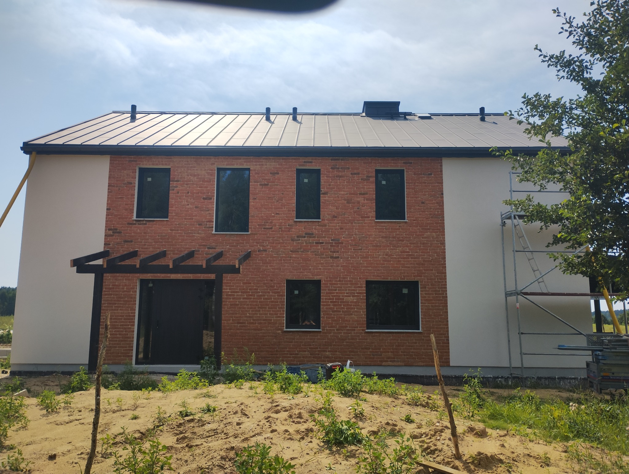 Dwukondygnacyjny dom jednorodzinny z elewacją częściowo wykończoną czerwoną cegłą i jasnym tynkiem, czarna stolarka okienna i drzwiowa, metalowy dach w kolorze grafitowym, widoczne rusztowanie przy...