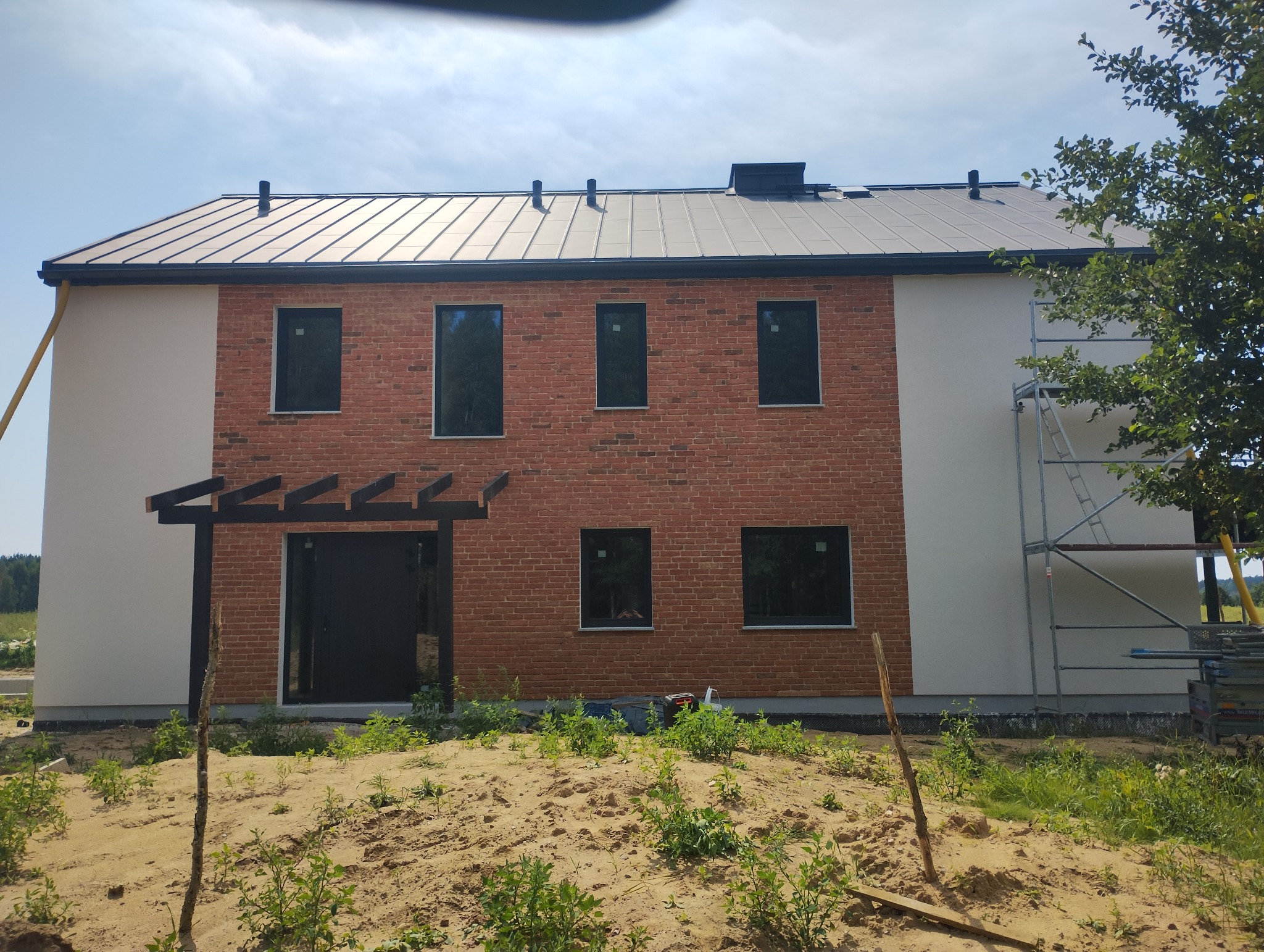Nowoczesny dom jednorodzinny z elewacją w kolorze cegły i jasnym tynkiem, widoczny fragment rusztowania i niezagospodarowany teren wokół budynku.