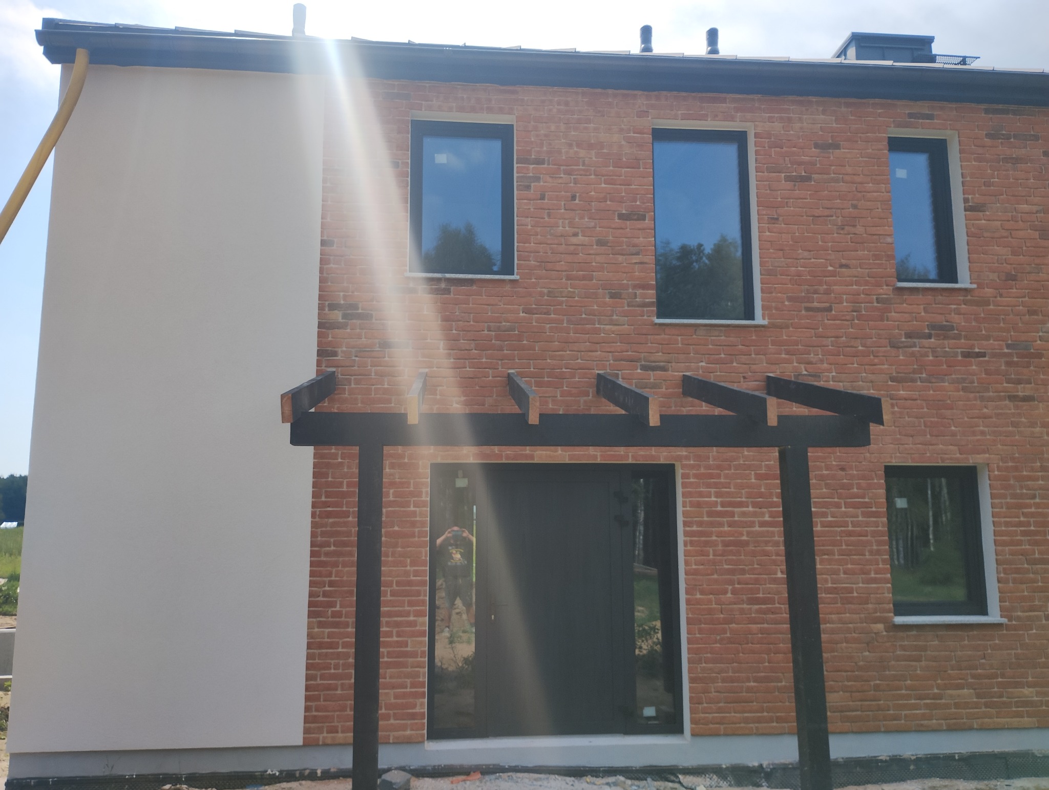 Dwukondygnacyjny dom z elewacją z czerwonej cegły i fragmentem ściany otynkowanej na biało, z czarnymi oknami i pergolą nad wejściem, w słoneczny dzień.