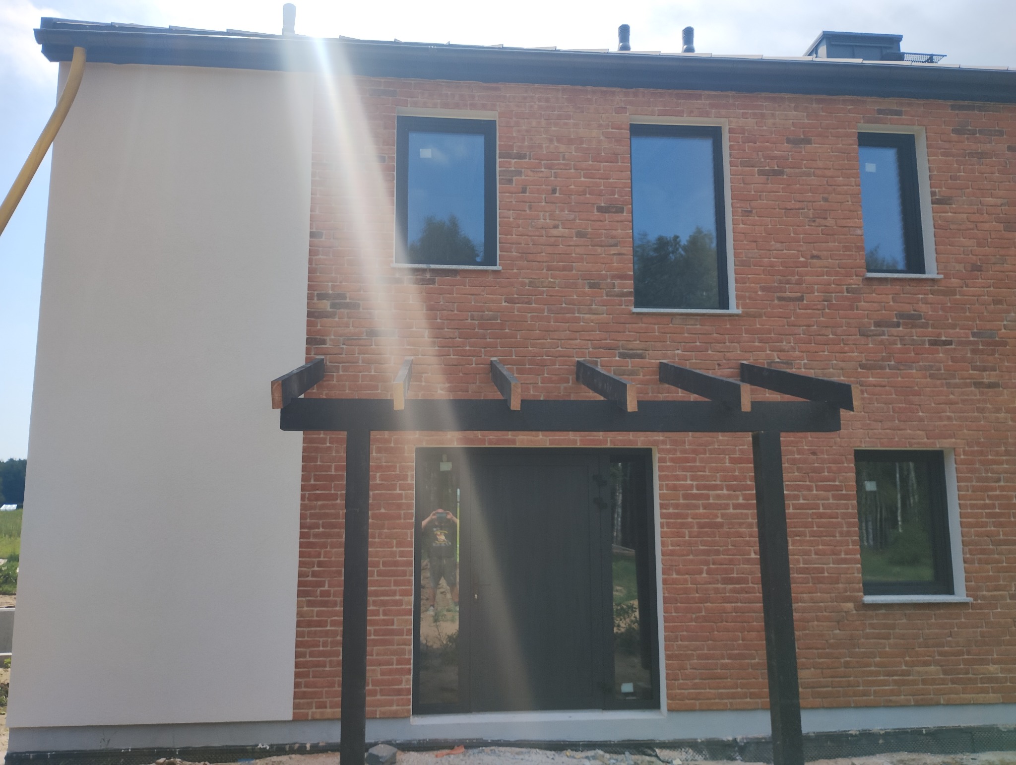 Dwukondygnacyjny dom z elewacją częściowo ocieploną i pomalowaną na biało, częściowo wykończoną czerwoną cegłą, z czarną stolarką okienną i drzwiową oraz drewnianą pergolą nad wejściem, w słoneczny...