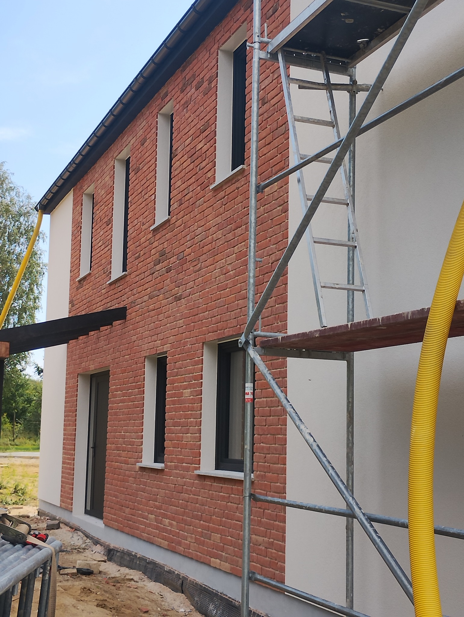 Dwukondygnacyjny budynek z częściowo wykończoną elewacją z cegły klinkierowej i jasnego tynku, widoczne rusztowanie z żółtym przewodem wentylacyjnym oraz przygotowany teren wokół budynku.