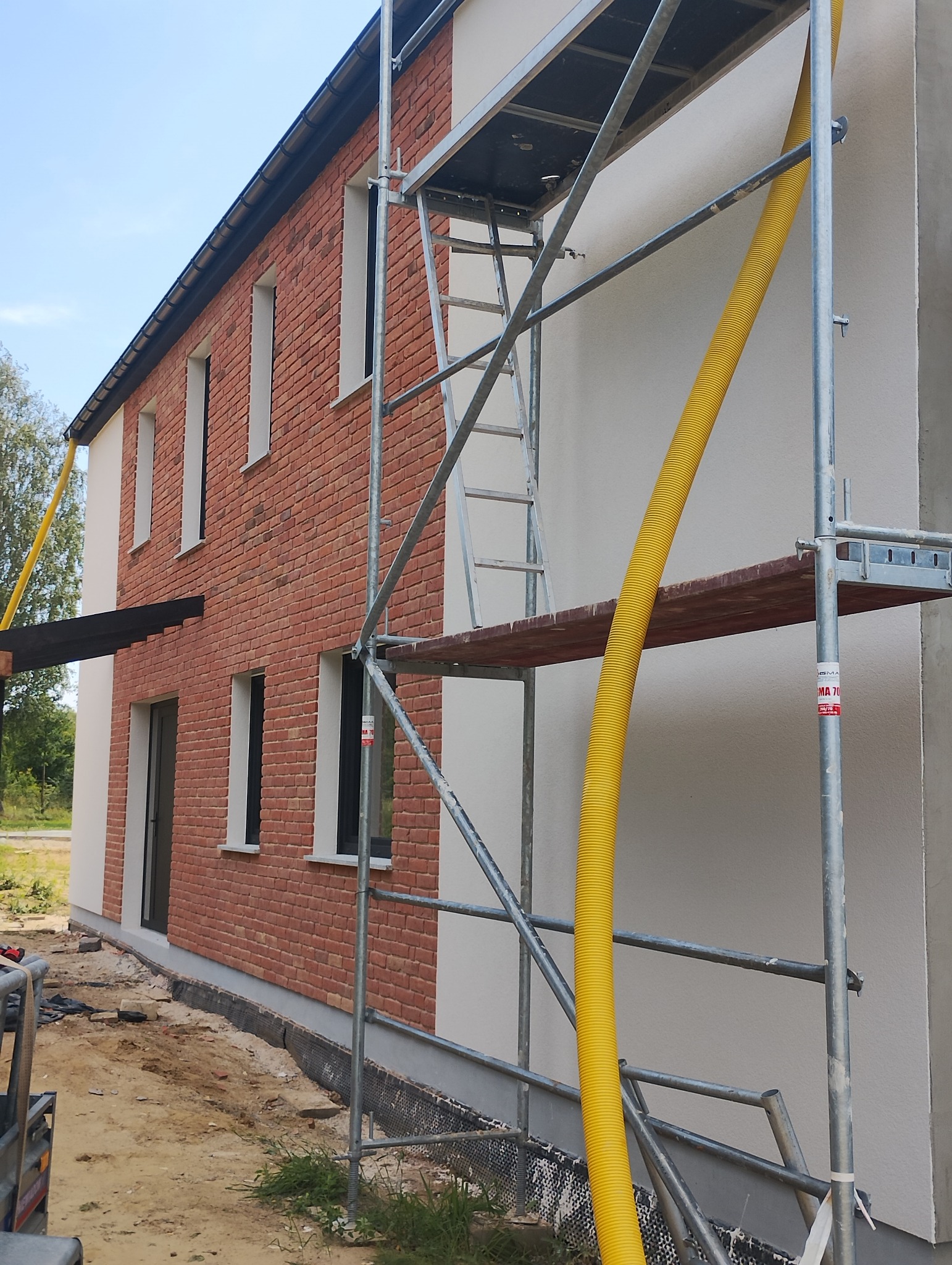 Dom z częściowo ocieploną elewacją w kolorze białym i ceglaną ścianą, widoczne rusztowanie z żółtym przewodem wentylacyjnym.