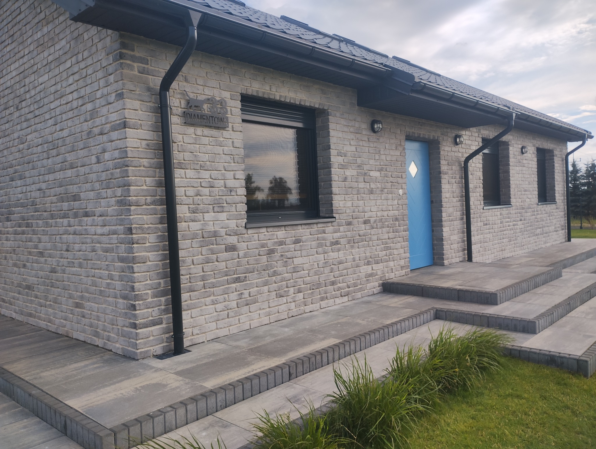 Elewacja z cegły klinkierowej w Dąbrównie. Szara cegła, czarna stolarka okienna i błękitne drzwi wejściowe. Widoczne schody i fragment trawnika.