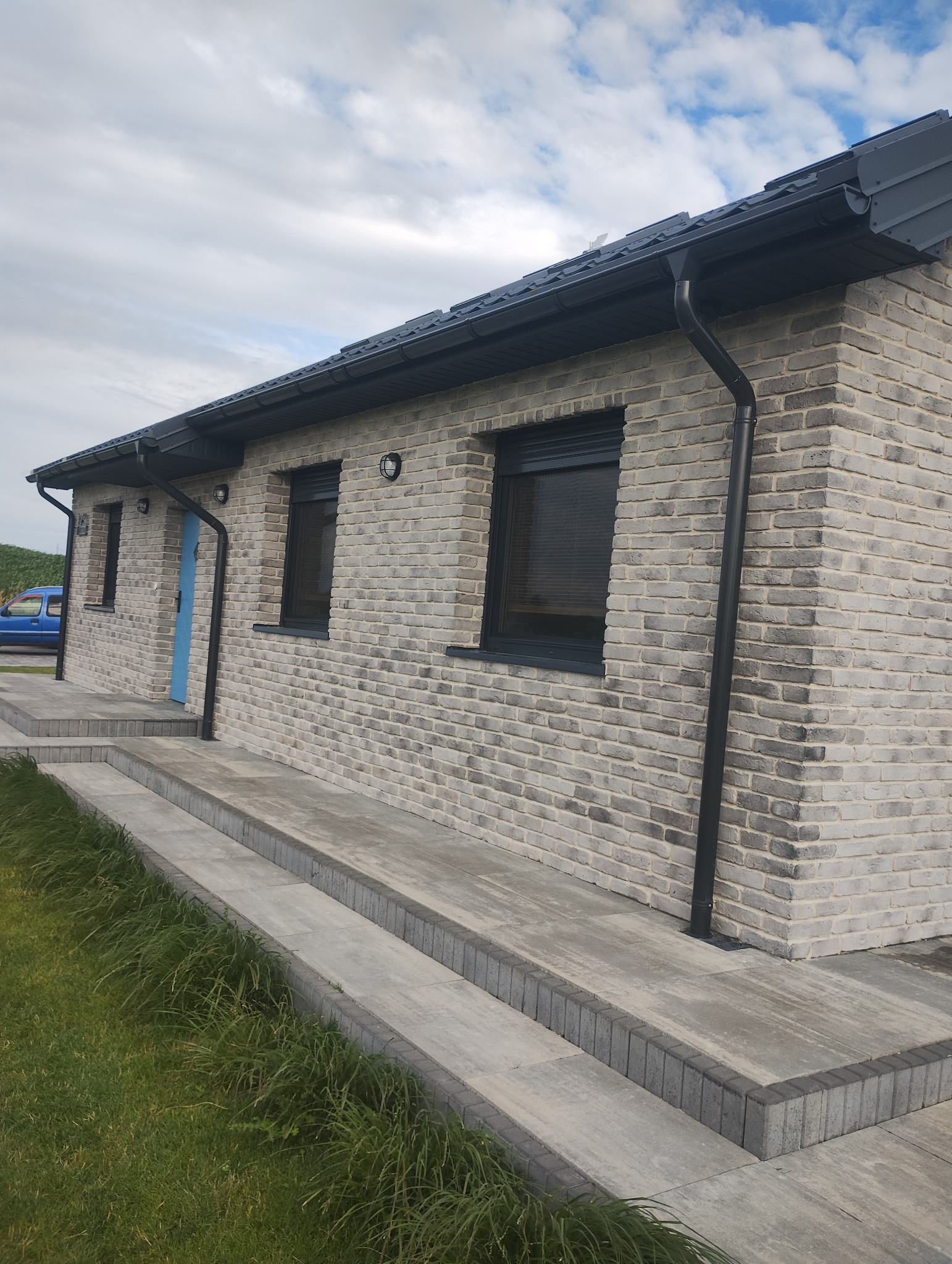 Nowoczesny dom z elewacją z szarej cegły klinkierowej, czarną stolarką okienną i grafitową rynną. Widoczne schody wejściowe i fragment trawnika. Błękitne niebo w tle.