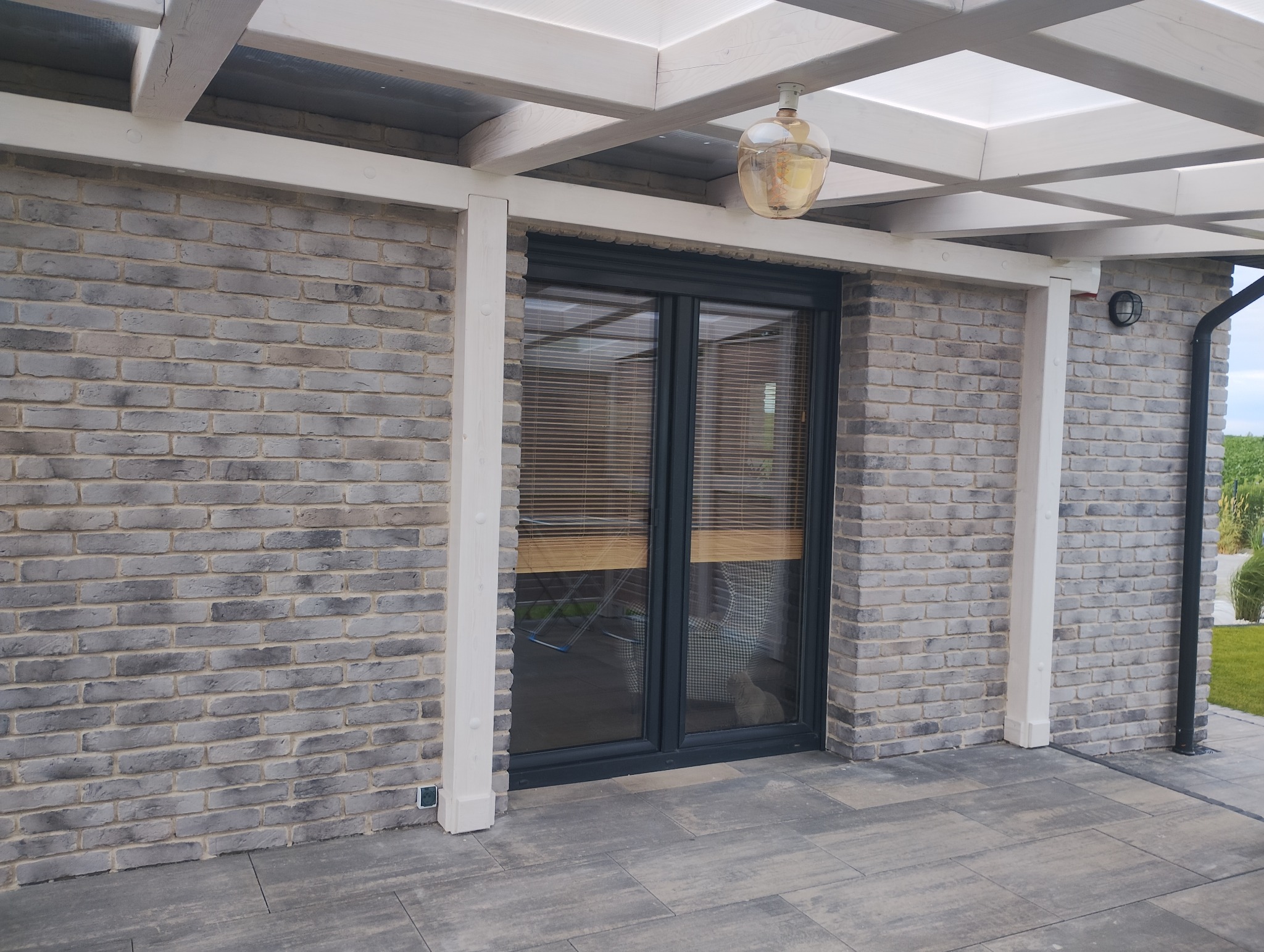 Szara ceglana elewacja domu z oknem i drewnianą pergolą, oświetlona wiszącą lampą. Widoczny fragment tarasu z szarych płyt.