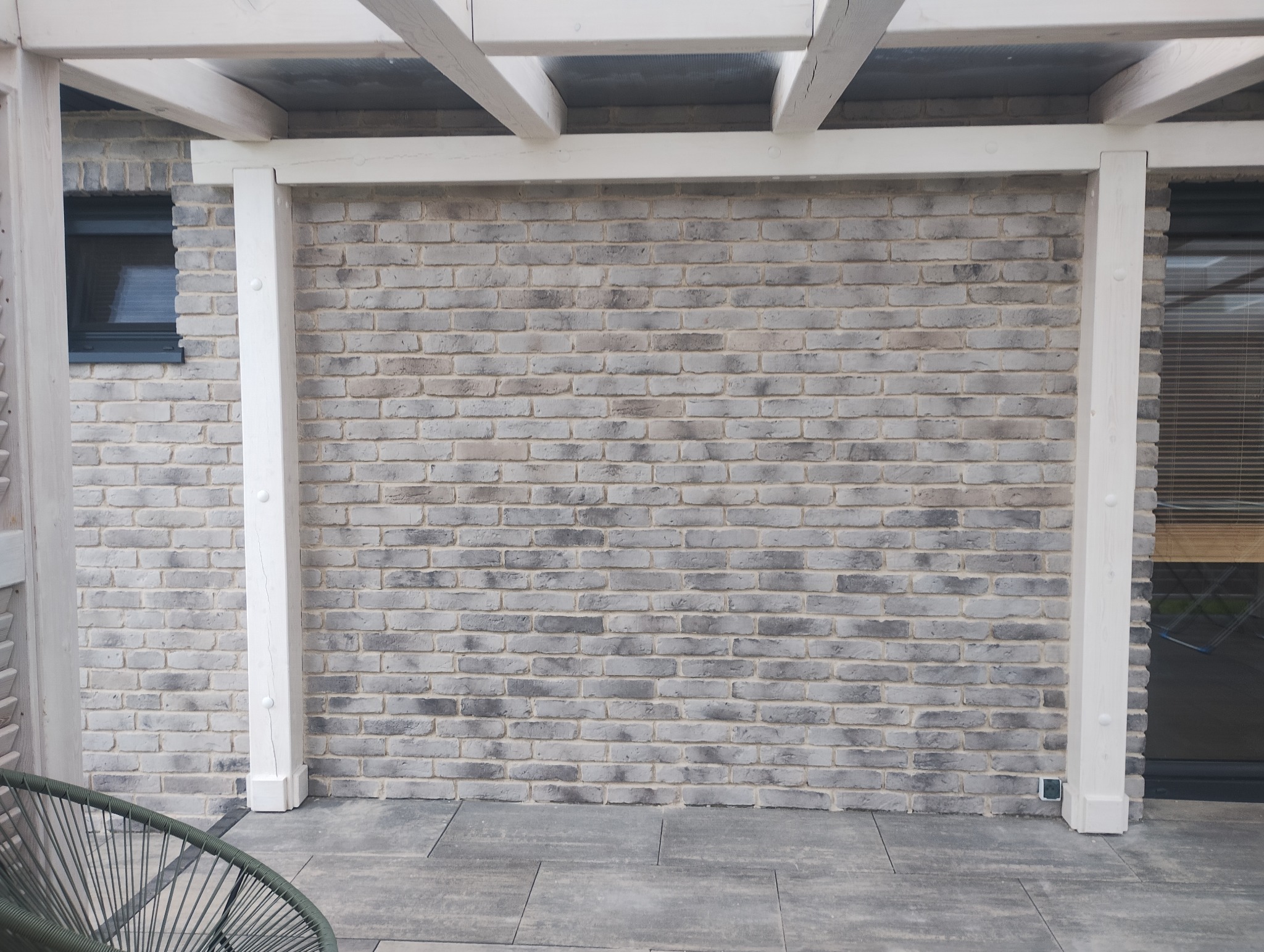 Murowana ściana z szarej cegły pod zadaszeniem z białych belek. Widoczne fragmenty okna, drzwi i krzesła. Szara, kamienna podłoga. Ujęcie frontalne.