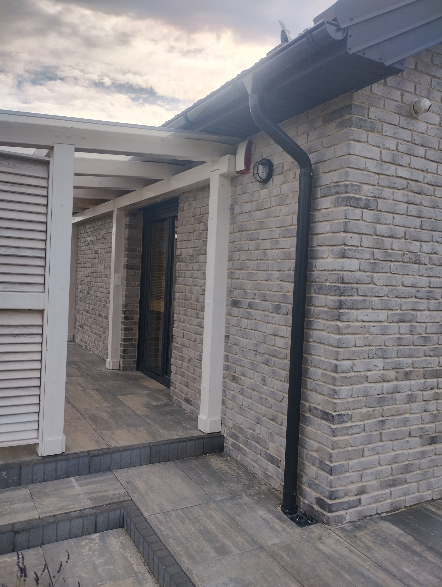 Szara ceglana elewacja domu z czarną rynną i białymi elementami konstrukcyjnymi werandy. Widoczne płytki chodnikowe i fragment nieba.