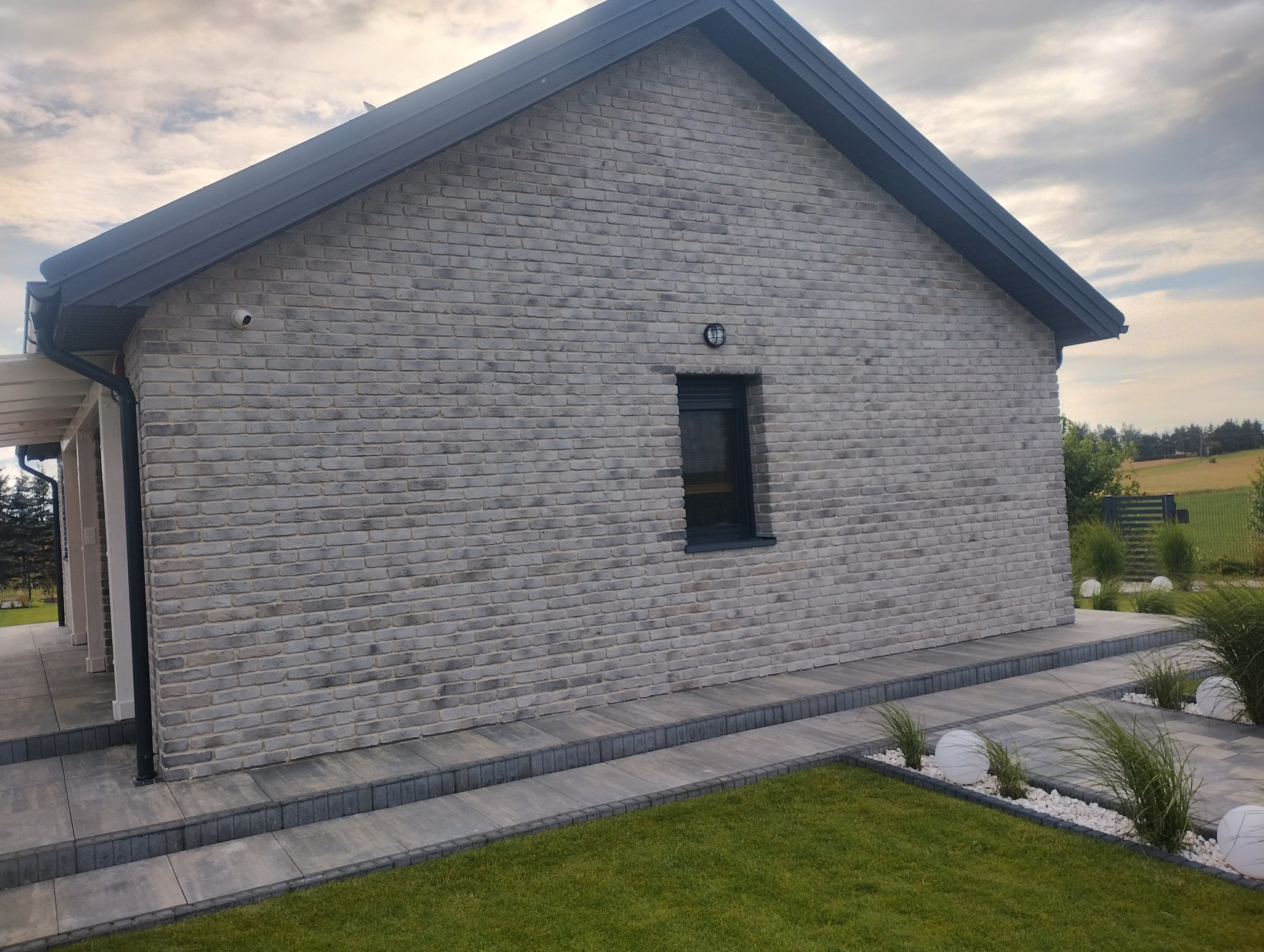 Elewacja domu z szarej cegły klinkierowej, widok z boku, z oknem i lampą zewnętrzną. Uwagę przykuwa staranne wykończenie i nowoczesny design.