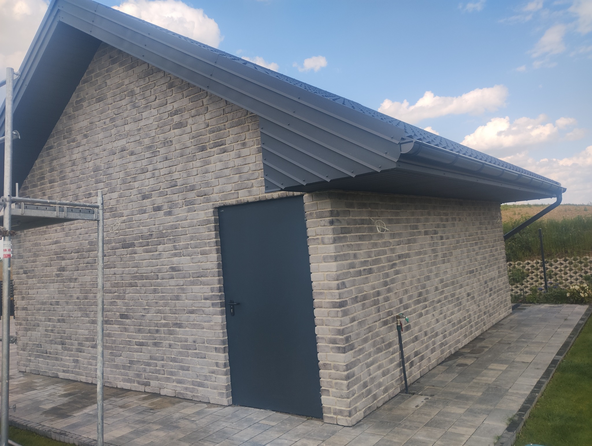 Murowana elewacja budynku z szarej cegły klinkierowej, z metalowym dachem i drzwiami, oraz widocznymi rusztowaniami. Całość w słoneczny dzień.