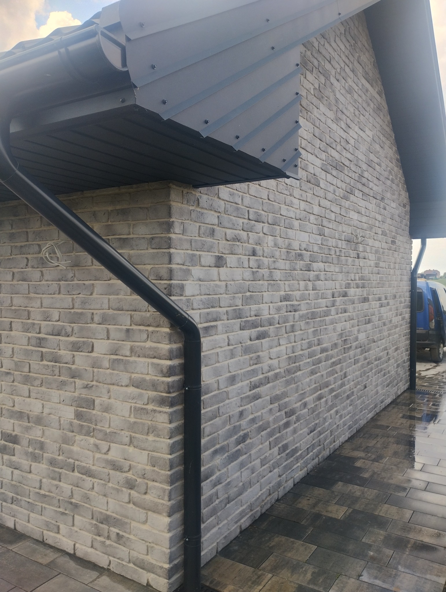 Szara elewacja z cegły klinkierowej, z widocznym orynnowaniem i fragmentem chodnika z kostki. Elewacja oświetlona naturalnym światłem.