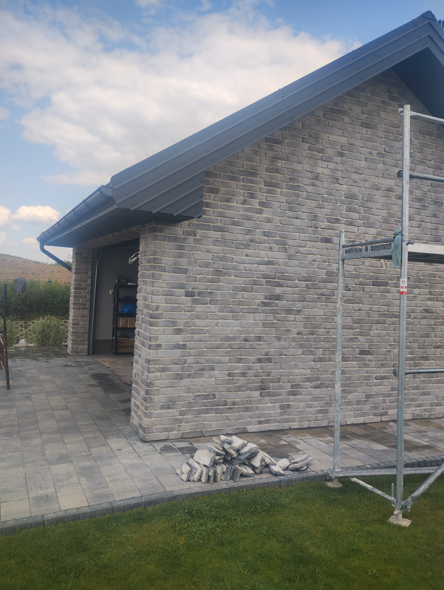 Murowana elewacja garażu z szarej cegły klinkierowej, widoczny fragment rusztowania i stos płytek elewacyjnych na kostce brukowej. Garaż z szarą blachodachówką.