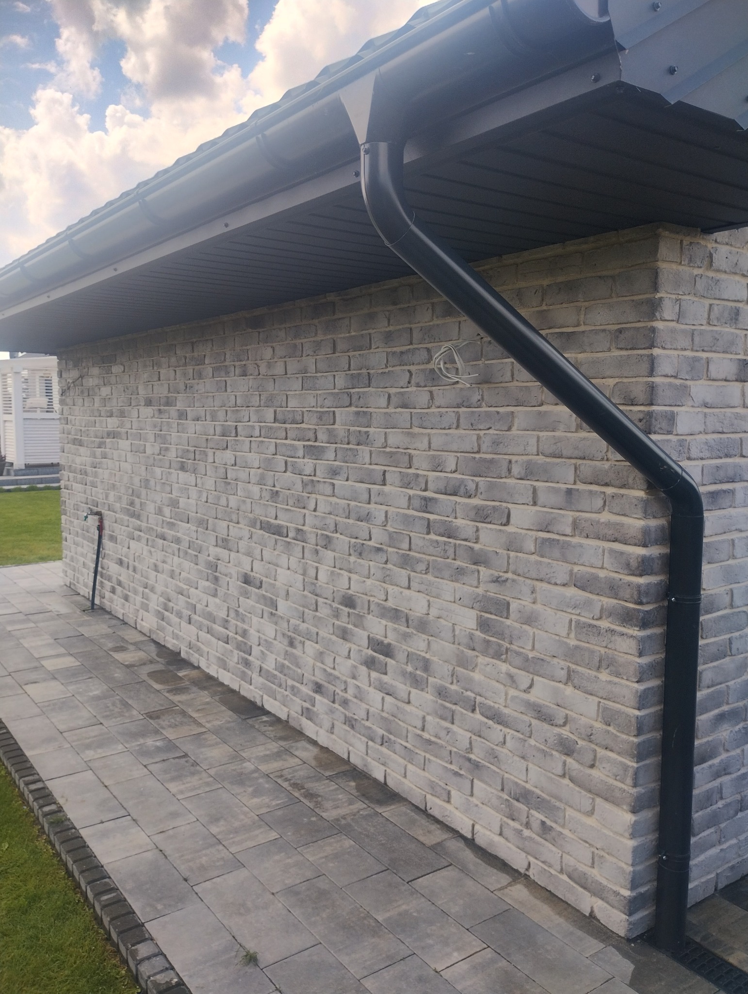 Elewacja budynku z szarej cegły klinkierowej, widoczny fragment orynnowania. Ułożona kostka brukowa. Na ścianie widoczne przewody elektryczne.