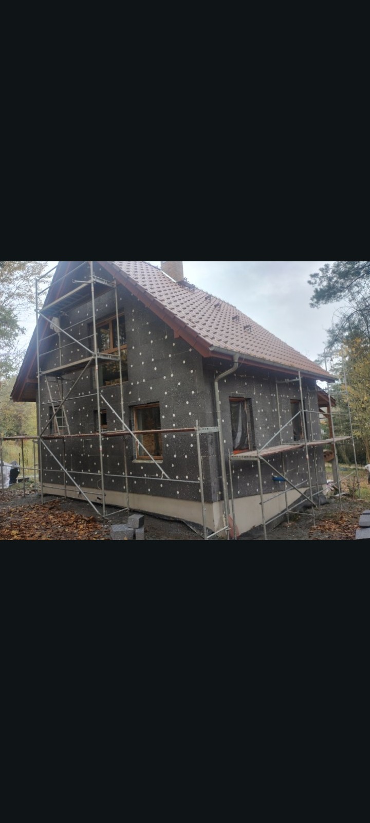 Dom jednorodzinny w trakcie ocieplania, widoczne rusztowania i styropian na elewacji. Ujęcie z boku, w tle drzewa i pochmurne niebo.