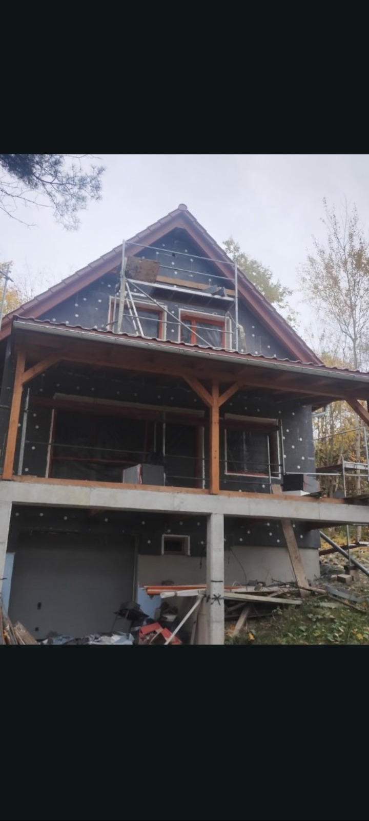 Dom w trakcie ocieplania ścian zewnętrznych, widoczne rusztowania i materiały izolacyjne. Częściowo wykończona elewacja z drewnianymi elementami.