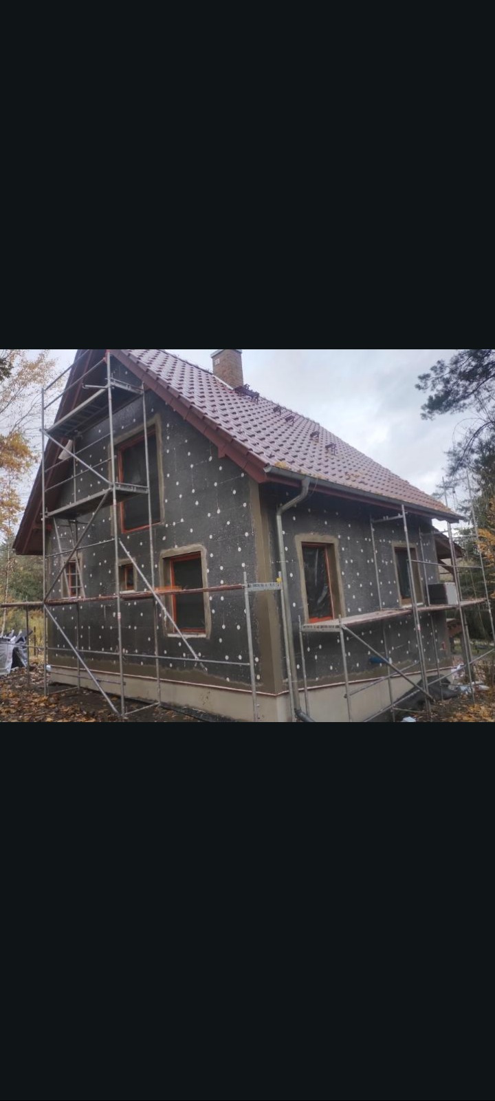 Dom w trakcie ocieplania, widoczne rusztowania i styropianowe kołki na szarej elewacji. Czerwony dach i jesienne drzewa w tle.