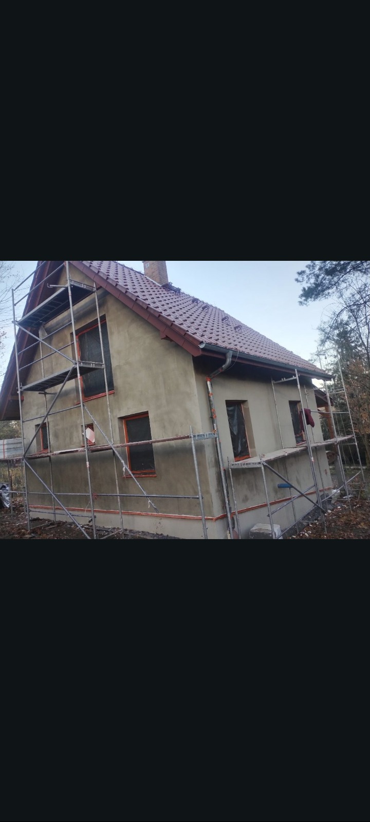 Dom jednorodzinny w trakcie ocieplania elewacji, widoczne rusztowania i surowa, szara elewacja. Okna zabezpieczone folią. Dach pokryty czerwoną dachówką.