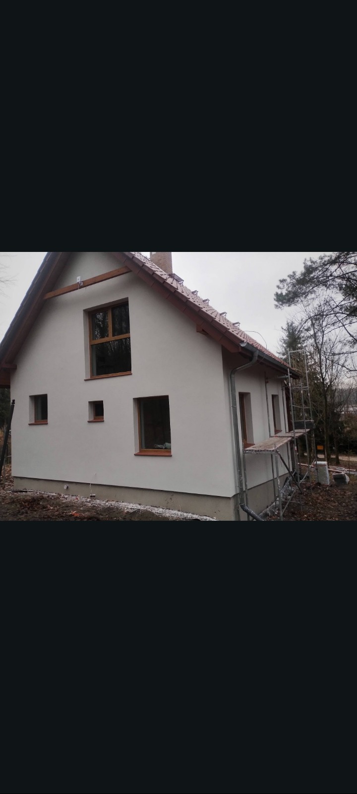Jasny, otynkowany dom jednorodzinny z drewnianymi ramami okiennymi, widziany z boku. Na ścianie widoczne rynny i rusztowanie. Tło stanowią drzewa.