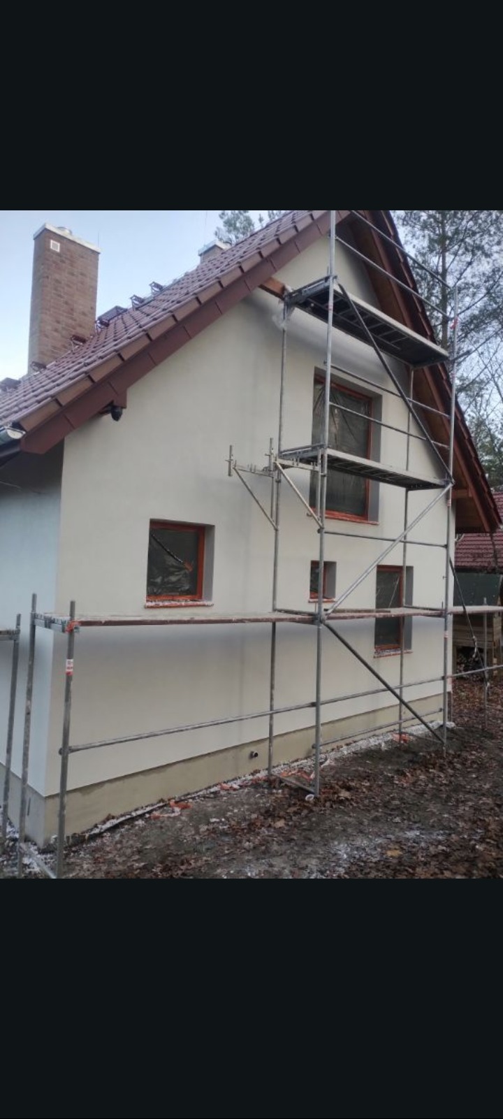 Dom jednorodzinny z jasną elewacją w trakcie prac, widoczne rusztowania. Okna zabezpieczone folią. Dach z brązowej dachówki, komin z cegły.