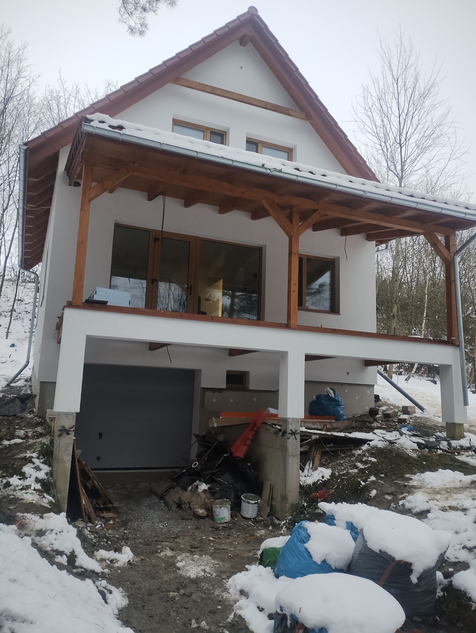 Dwukondygnacyjny dom z białymi ścianami i drewnianą konstrukcją werandy, otoczony zimowym krajobrazem z resztkami śniegu i widocznymi śladami prac budowlanych.