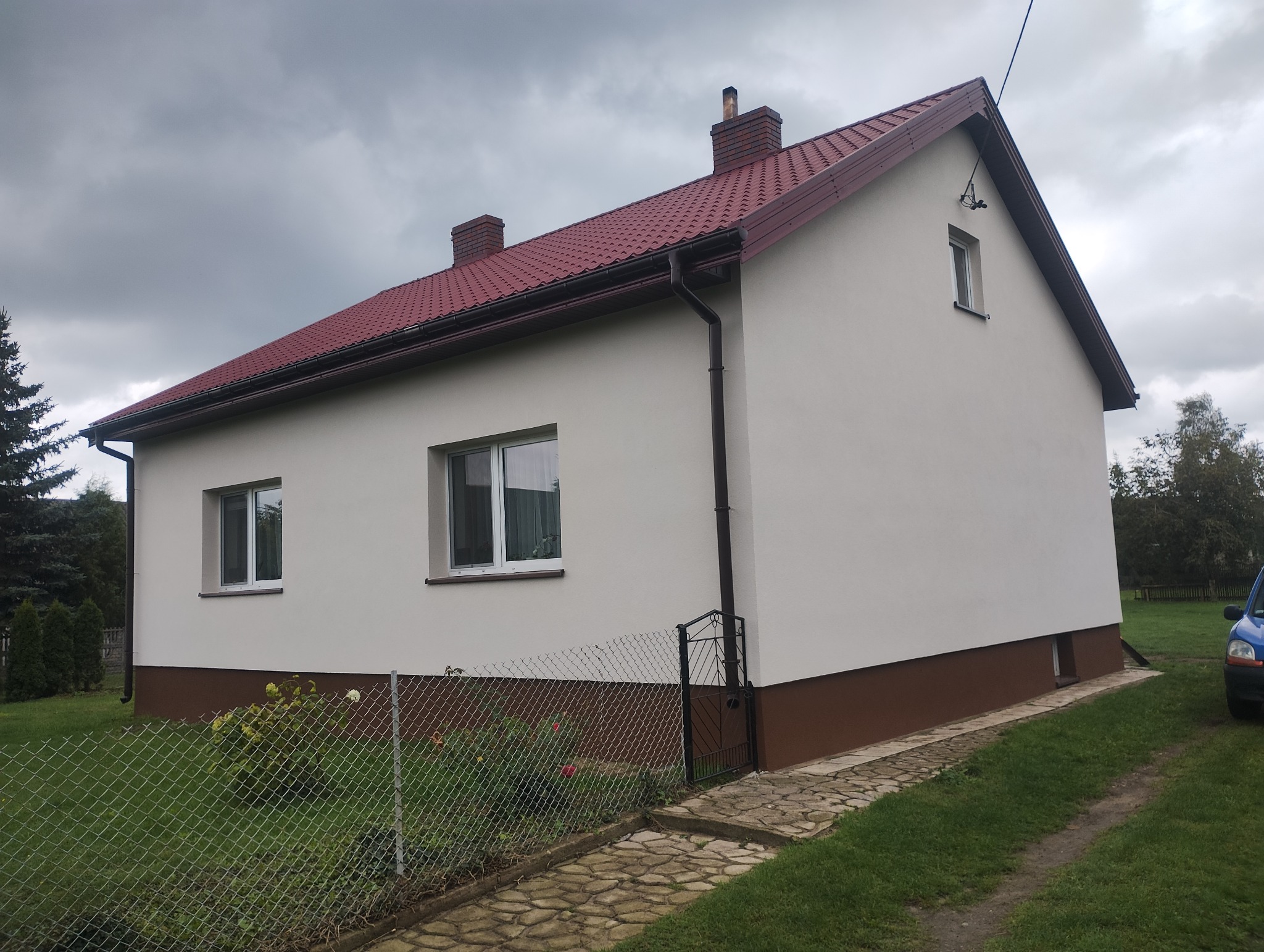 Dom jednorodzinny po remoncie elewacji, z nową, jasną elewacją i ciemnobrązowym cokołem. Czerwony dach, widoczne okna i fragment ogrodzenia.