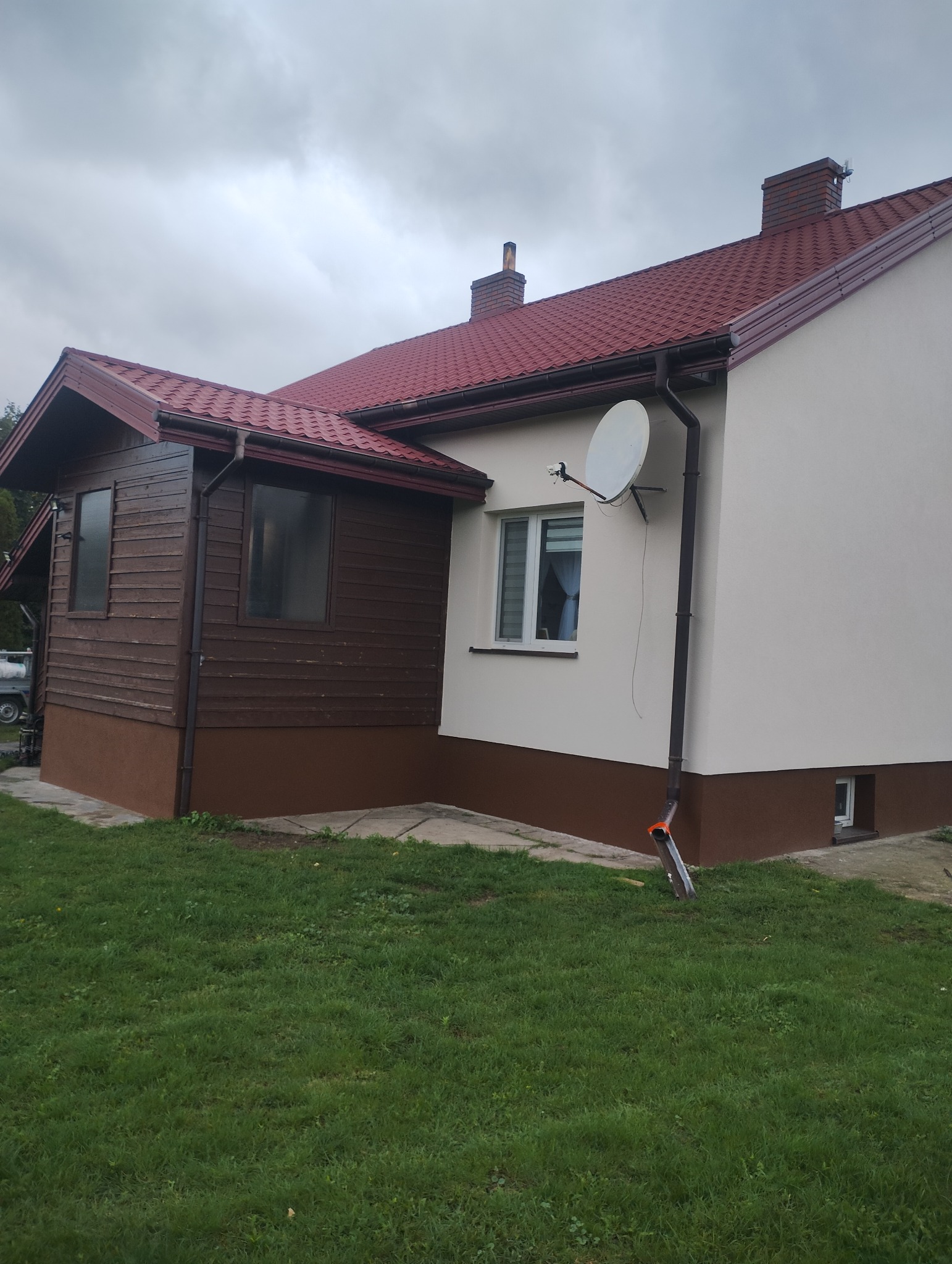 Dom jednorodzinny po remoncie elewacji, z widoczną drewnianą dobudówką, białą elewacją, czerwoną dachówką i anteną satelitarną. Widok z poziomu trawnika.