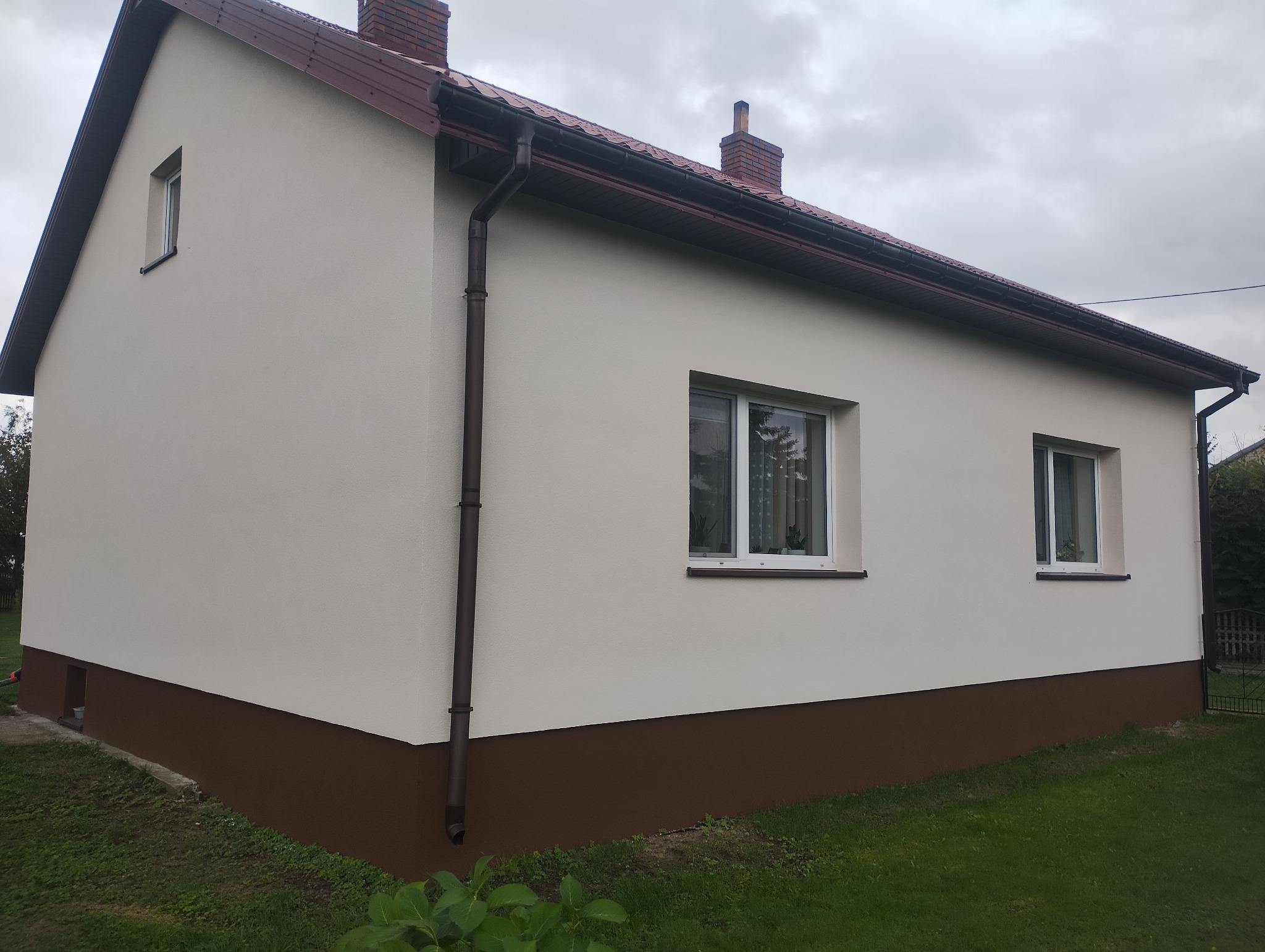 Jasna elewacja domu jednorodzinnego z brązowym cokołem i orynnowaniem, widok z boku na tle zielonego trawnika i pochmurnego nieba. Detal: widoczne dwa okna.