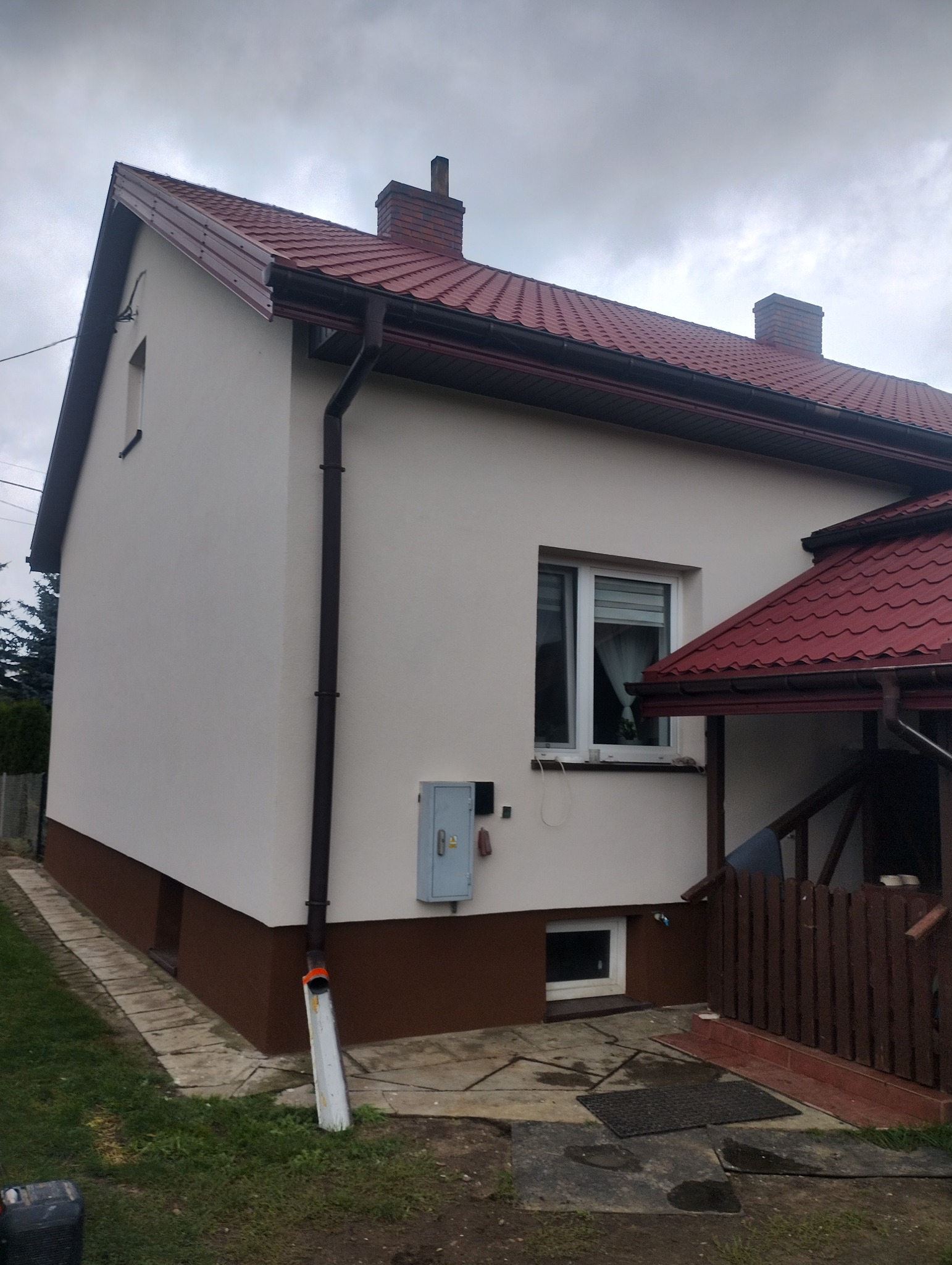 Dom po renowacji elewacji z nowym, brązowym orynnowaniem i pomalowaną na brązowo dolną częścią. Widoczne okno z roletami i fragment dachu ganku.