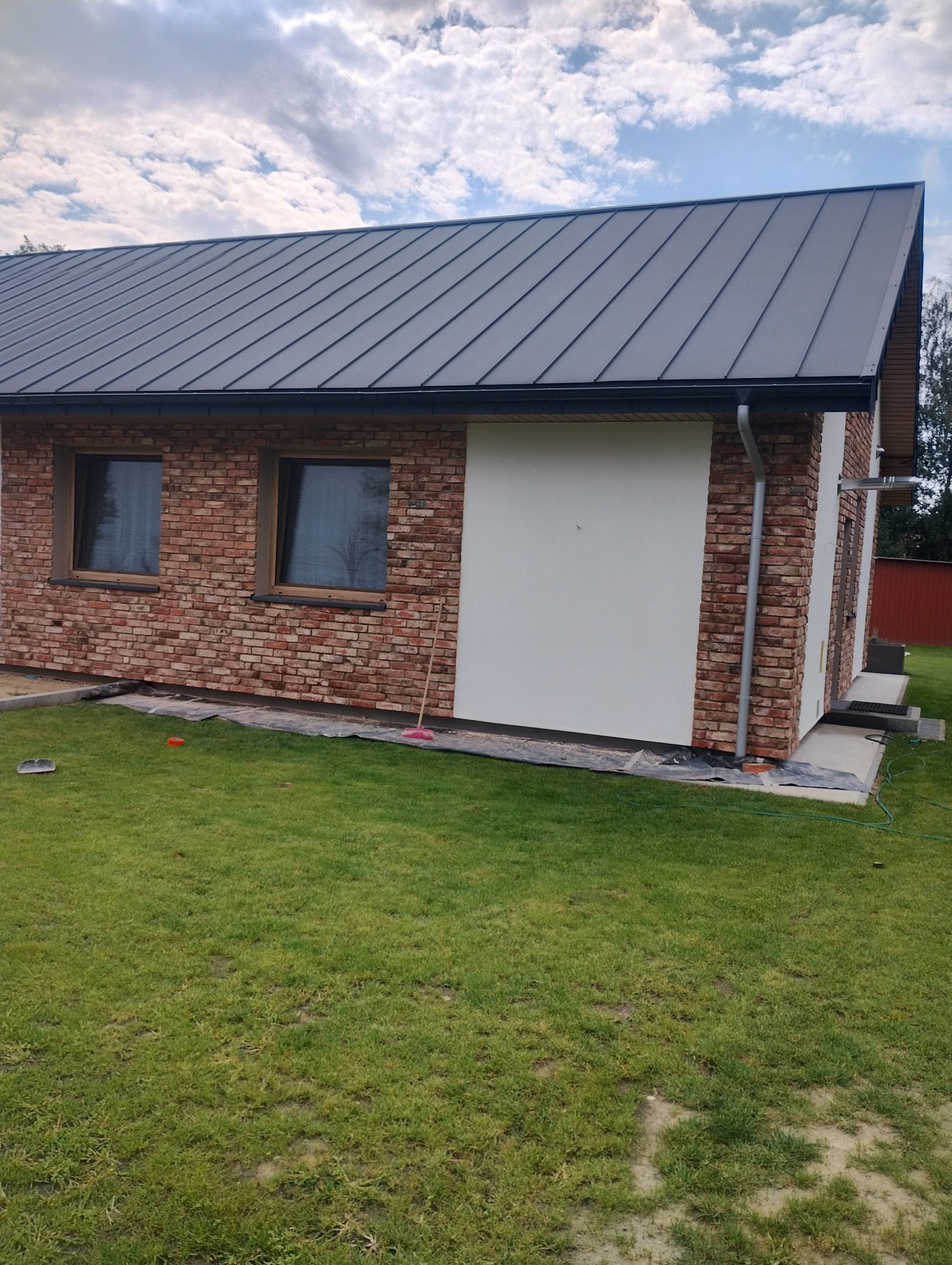 Dom z elewacją z cegły klinkierowej i fragmentem otynkowanej ściany. Widoczny nowy, grafitowy dach i rynna. Na trawniku leżą narzędzia i folia zabezpieczająca.