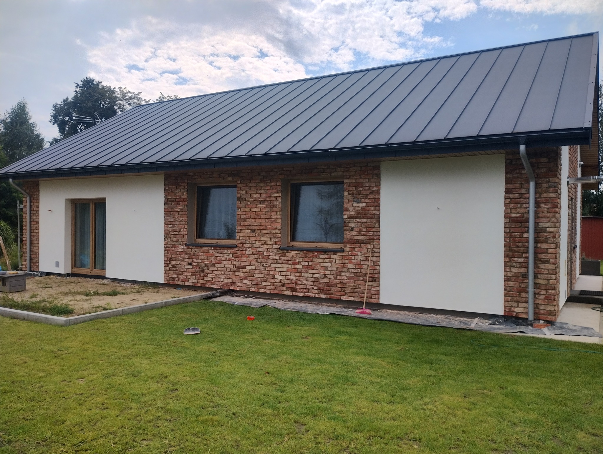 Parterowy dom z elewacją łączącą biały tynk i cegłę klinkierową, grafitowy dach, okna w drewnianych ramach. Widok z trawnika w słoneczny dzień.