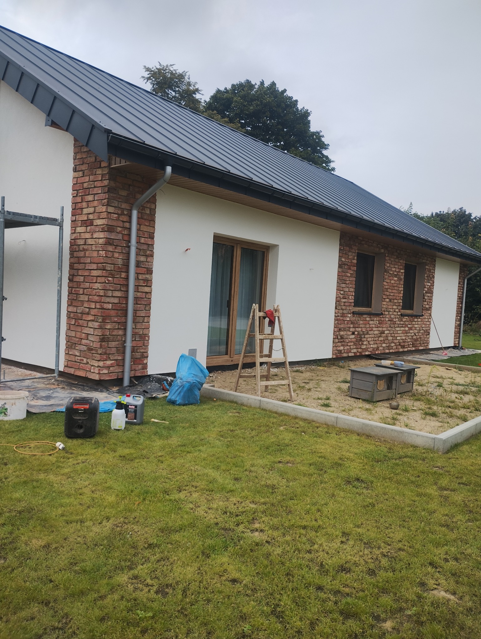 Dom jednorodzinny z fragmentami elewacji wykończonymi czerwoną cegłą, częściowo otynkowany na biało. Widoczne narzędzia i drabina sugerują trwające prace wykończeniowe.