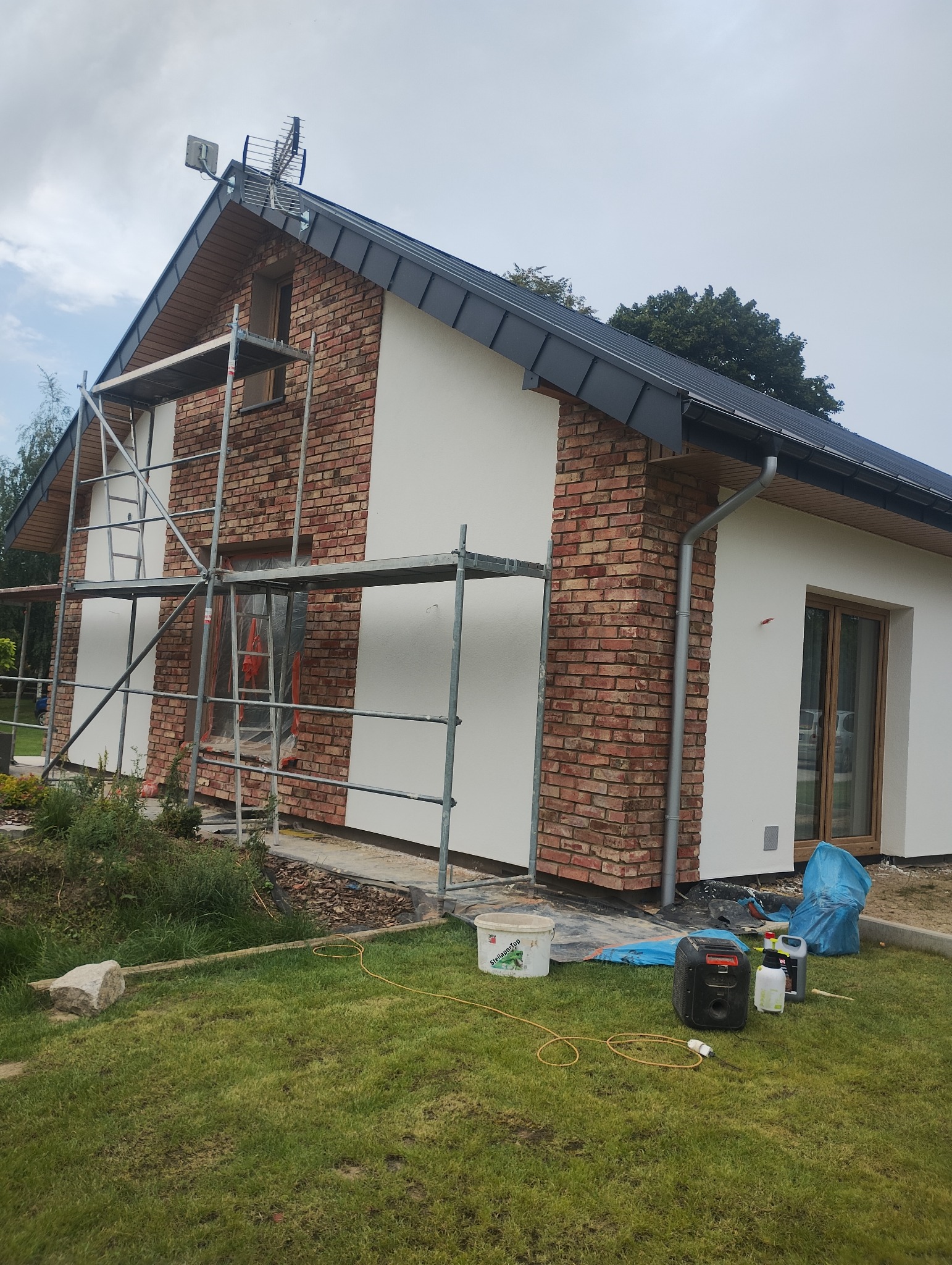 Dom z elewacją w trakcie prac: fragmenty ceglanej ściany i białe, gładkie powierzchnie. Widoczne rusztowanie, narzędzia malarskie i akcesoria na trawniku.