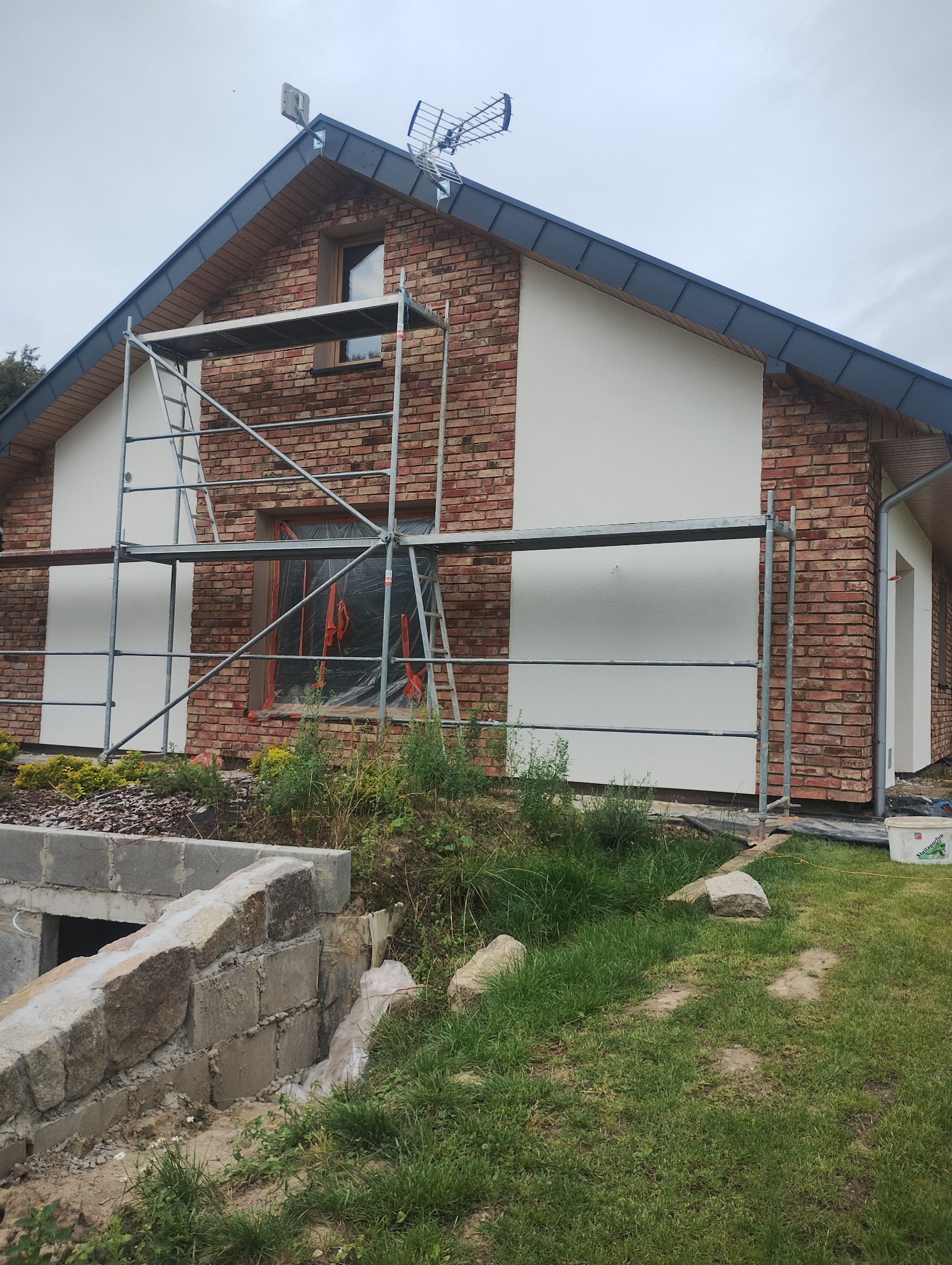 Dom z ceglaną elewacją w trakcie prac: fragmenty otynkowane na biało, rusztowanie, zabezpieczone okno. Widoczny fragment fundamentu i trawnik.