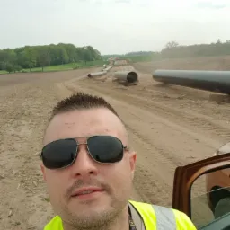 Mężczyzna w okularach i żółtej kamizelce odblaskowej robi selfie na tle rurociągu w trakcie budowy, widoczne fragmenty samochodu.