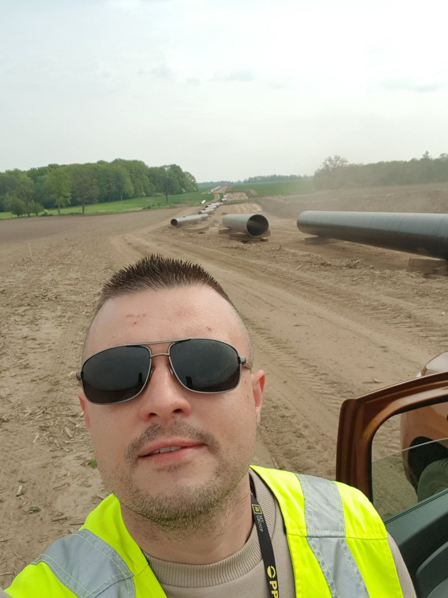 Mężczyzna w okularach i żółtej kamizelce odblaskowej robi selfie na tle rurociągu w trakcie budowy, widoczne fragmenty samochodu.