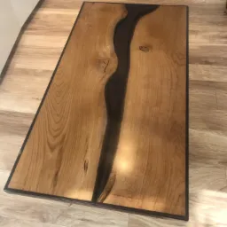 Blat stołu z litego drewna z żywicą epoksydową tworzącą wzór rzeki, wykończony ciemną ramą, leżący na panelach podłogowych.