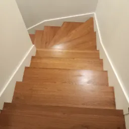 Widok z g&oacute;ry na drewniane schody z oświetleniem LED wzdłuż ściany, tworzące spiralę w nowoczesnym wnętrzu.