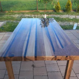Stół z blatem z drewna egzotycznego z żywicą epoksydową, widoczne połączenia stolarskie, na zewnątrz, w tle trawa i drzewa.