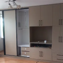 Beżowa szafa wnękowa z przesuwnymi drzwiami i częścią RTV, zlokalizowana w Nowym Korczynie, z widocznymi niedoskonałościami wykończenia i kubkiem na półce.