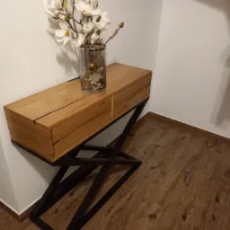 Drewniana konsola z dwiema szufladami na metalowym, geometrycznym stelażu, udekorowana wazonem z białymi kwiatami i ozdobami, stoi przy białej ścianie na podłodze wyłożonej panelami imitującymi...