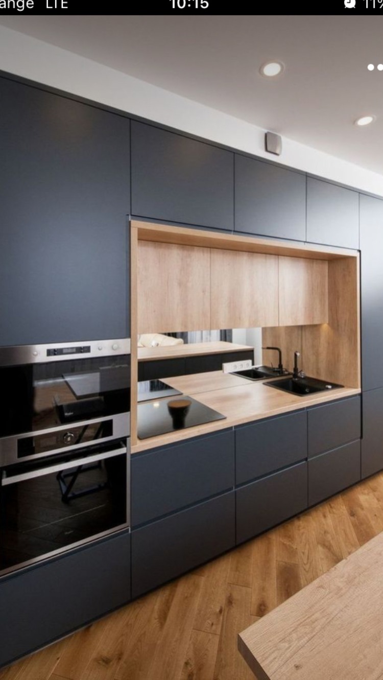 Nowoczesna kuchnia z ciemnymi, matowymi frontami szafek i drewnianym akcentem w otwartej przestrzeni nad blatem, wyposażona w piekarnik i płytę grzewczą, z drewnianą podłogą w jodełkę.